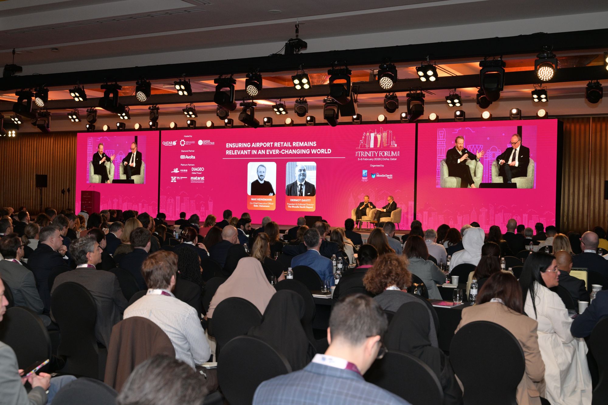 Conference hall with a pink theater screen showing two speakers seated on stage and an audience in foreground