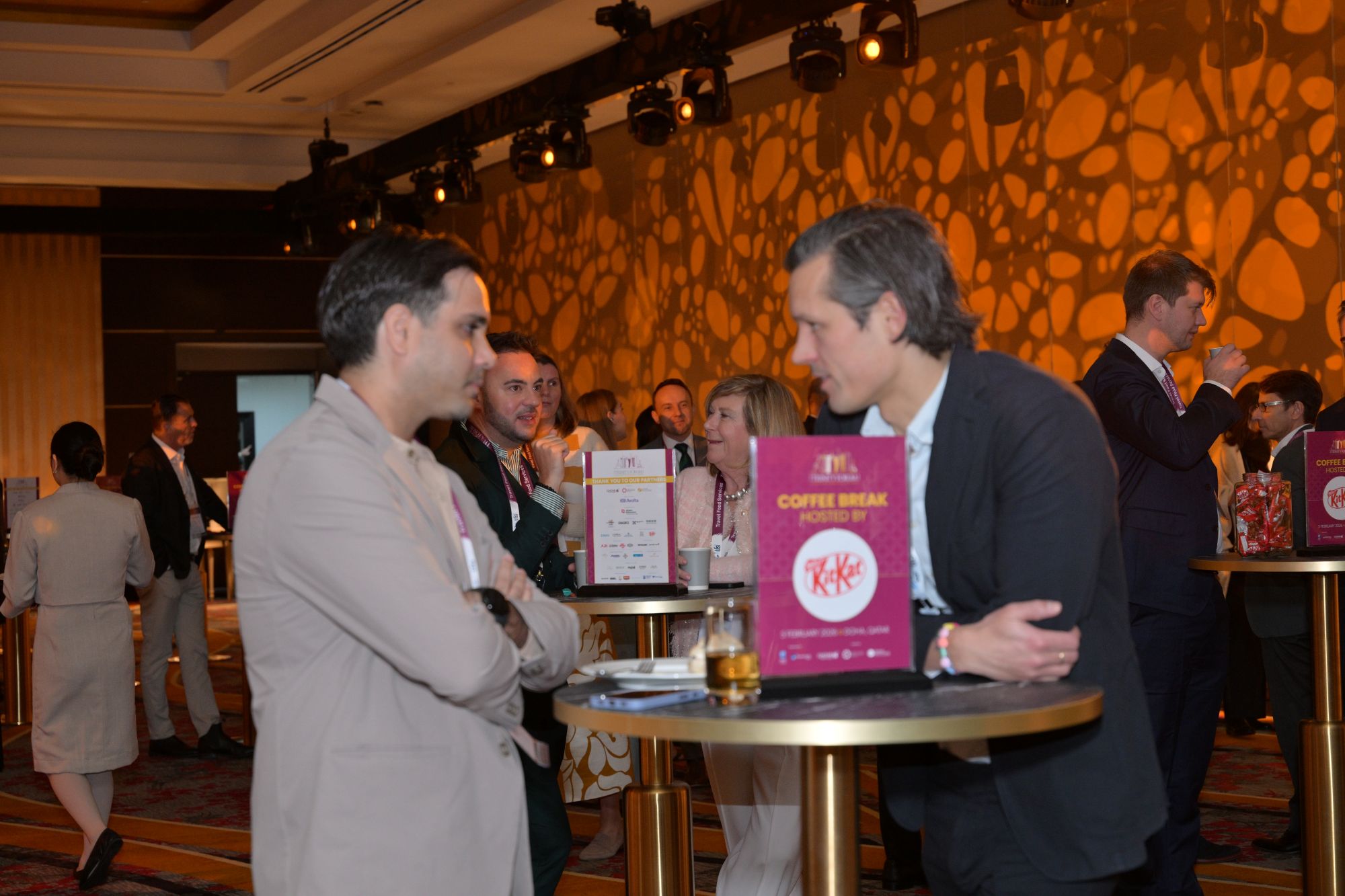 Professionals in business attire mingle around round high tables at a conference lounge, warm lighting and a pink Coffee Break sign in the foreground.