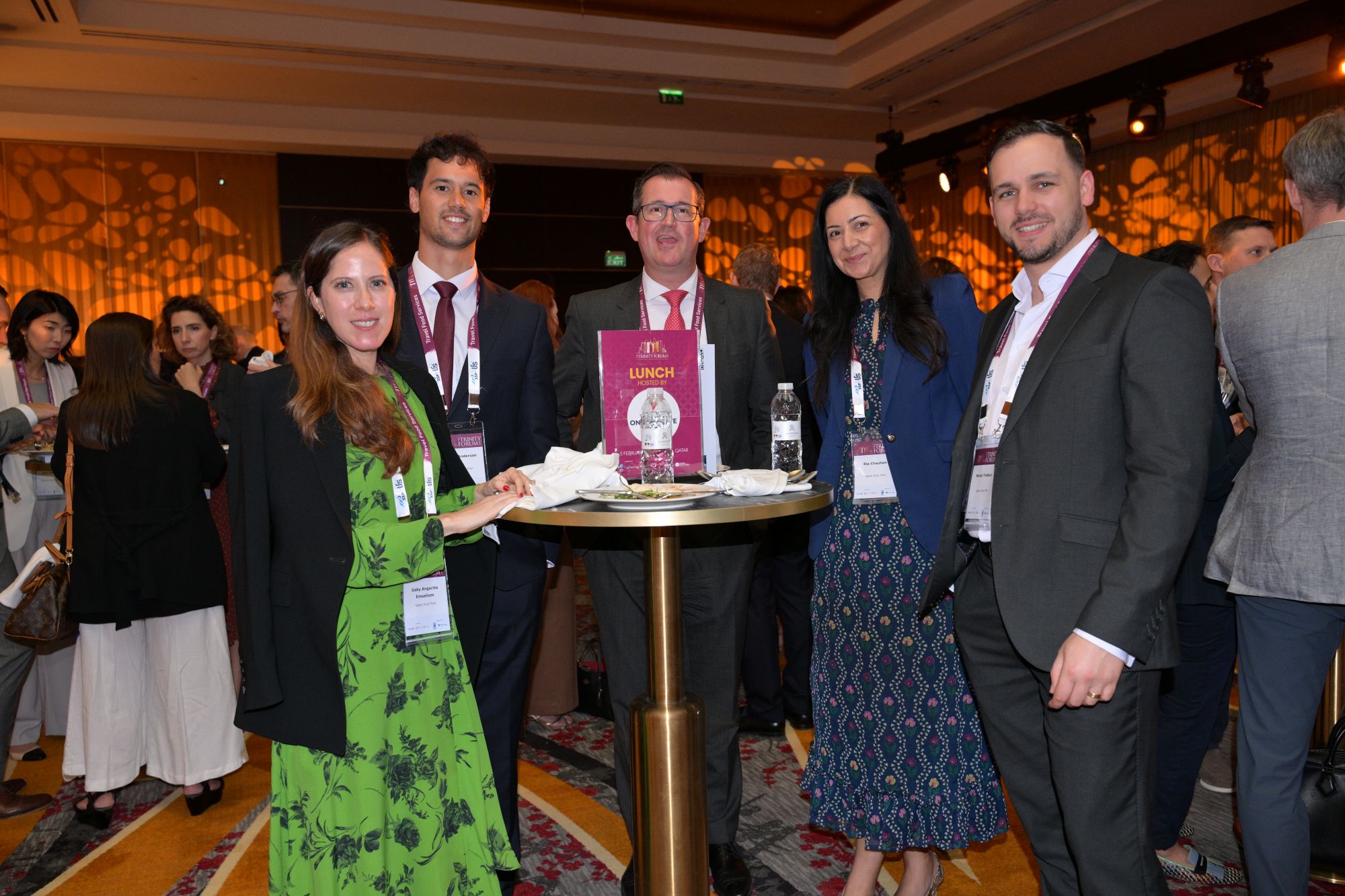 Group of professionally dressed people around a tall round table at a networking event, smiling at the camera in a conference venue
