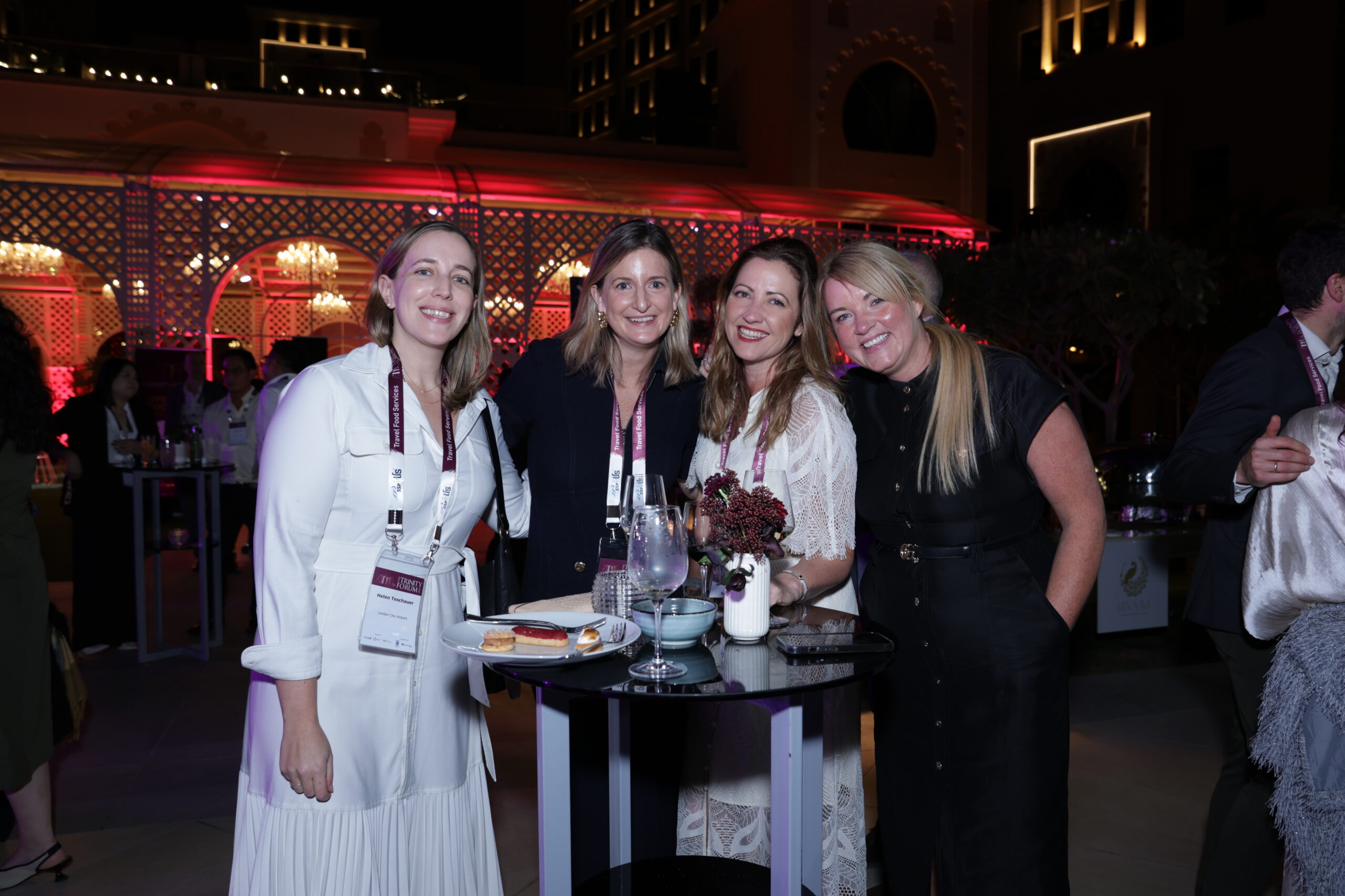 Four women in elegant outfits smile at a nighttime cocktail event, standing by a high table with drinks and snacks.