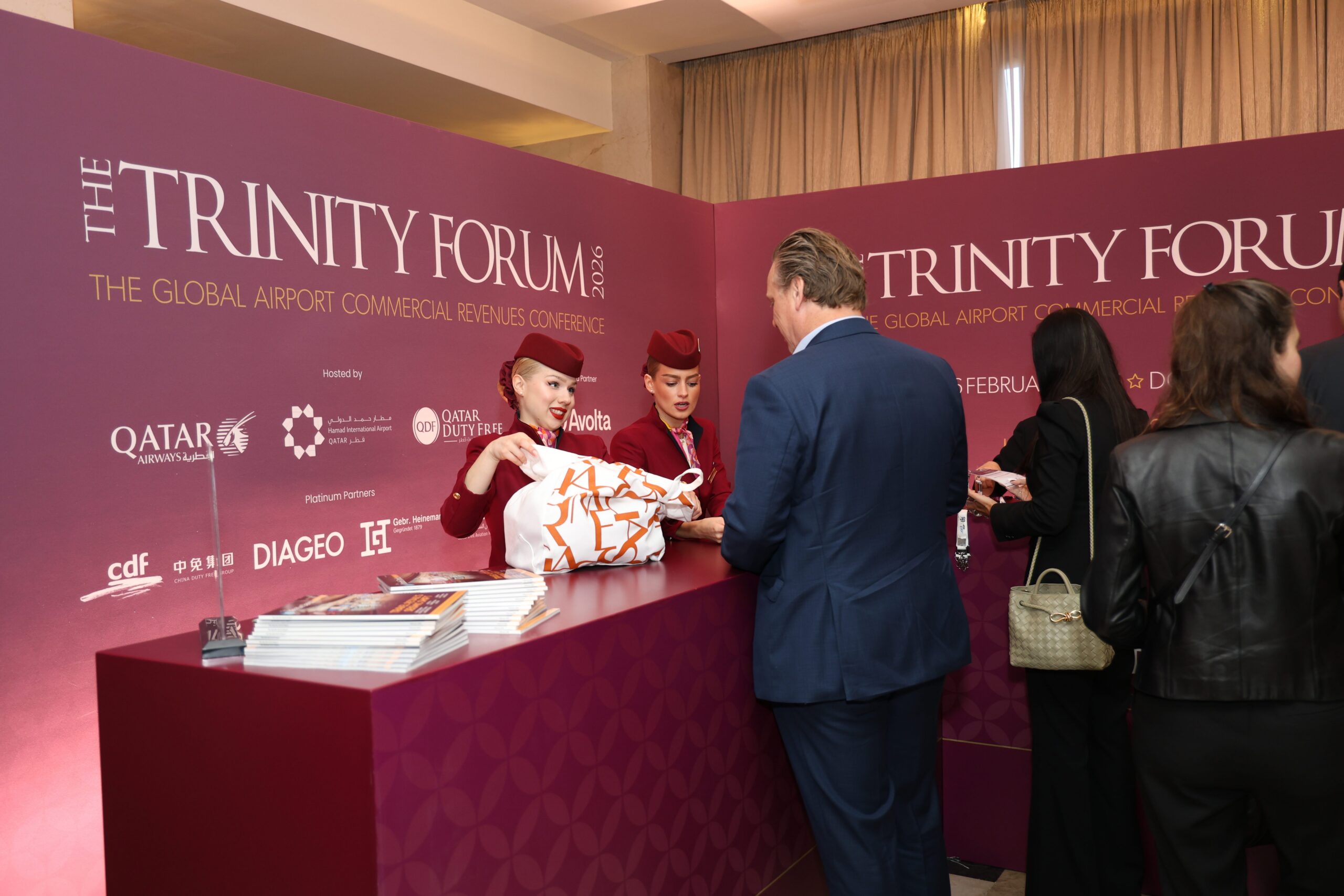 Registration desk at a purple Trinity Forum conference, two uniformed staff handing a tote to a man in a blue suit.
