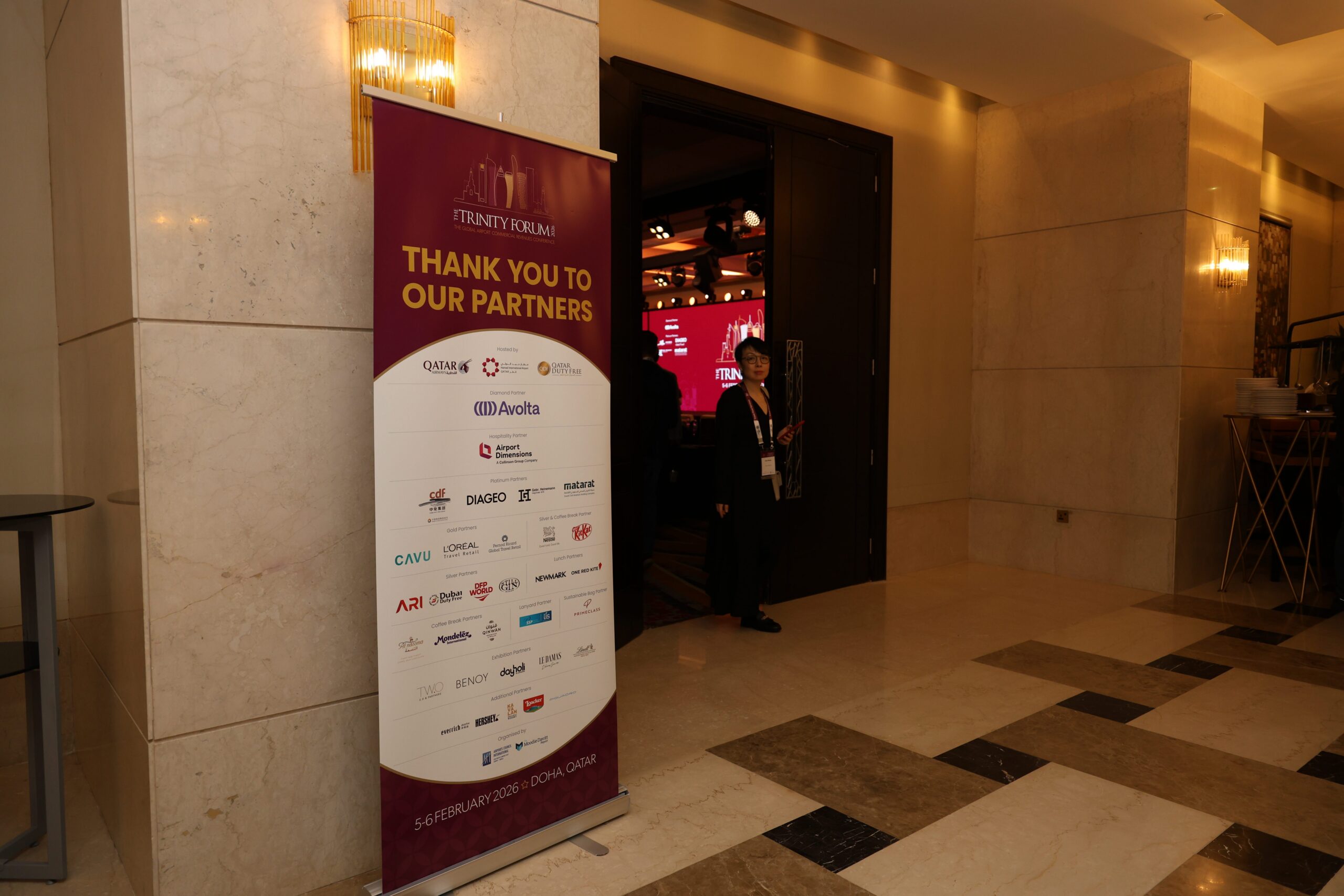 Banner reading 'Thank you to our partners' in a burgundy display stand at an event entrance, with a person standing near an open door in a marble lobby.