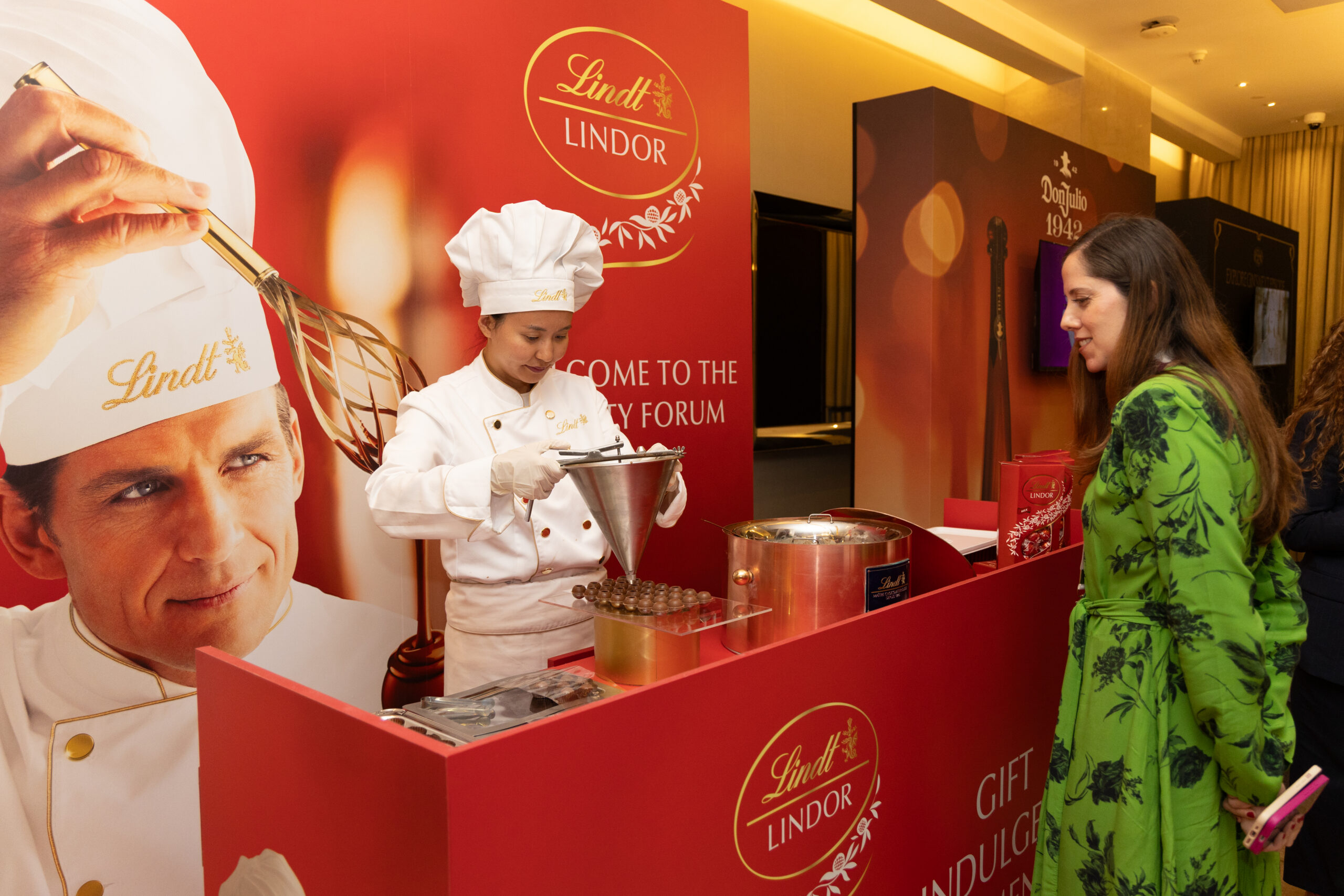 Lindt promotional booth with a chef in white pouring chocolate into a funnel while a visitor watches, red Lindt banners in the background.