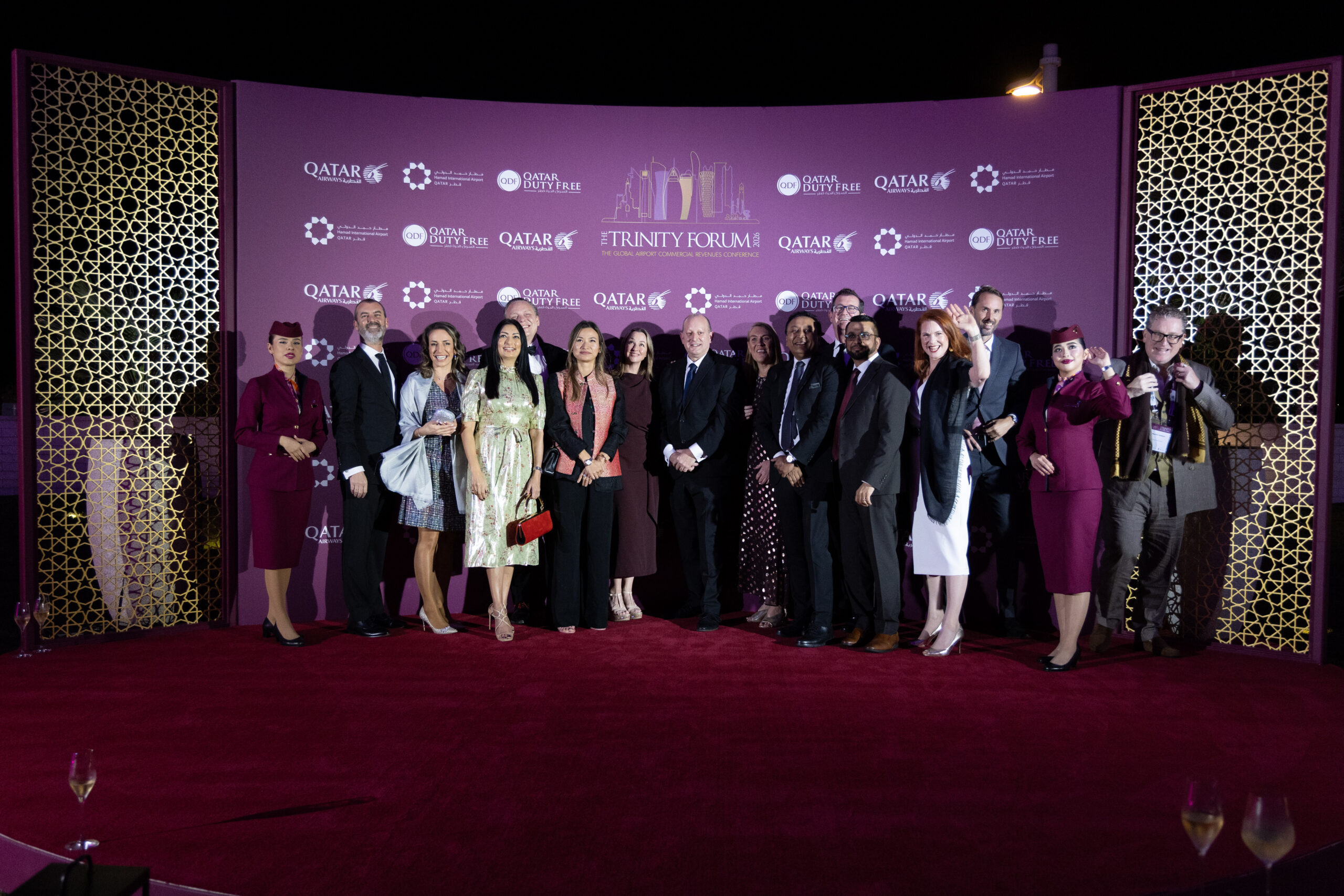 Group of professionally dressed people posing on a red carpet in front of a purple backdrop with logos, flanked by gold lattice panels at a formal event.